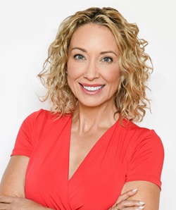The image features a medium-light-skinned woman with shoulder-length curly blonde hair. She has a warm smile and is wearing a red V-neck top. The woman's makeup is natural, complementing her clear complexion, and she has light pink lipstick. Her eyes are bright and friendly, and she is looking directly at the camera. The background is white and unadorned, providing a stark contrast that highlights her features.