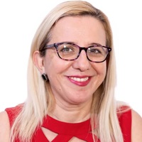 This is a headshot of Maja Zelihic against a white backdrop. She has light skin and straight blonde hair that hangs below her shoulders. Her hair is parted on her left side and tucked behind her right ear. She has medium brown eyes and an inviting smile. She wears purple tortoise shell glasses and large black stone earrings. Her lipstick is a light raspberry red and matches her red sleeveless blouse which has three triangle cutouts along her collarbone.